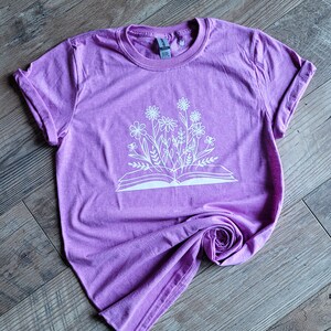 Peut inclure: Un t-shirt violet avec un motif blanc d'un livre ouvert avec des fleurs qui en sortent.