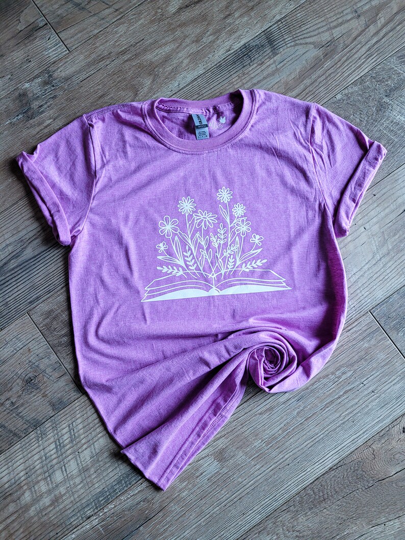 Peut inclure: Un t-shirt violet avec un motif blanc d'un livre ouvert avec des fleurs qui en sortent.