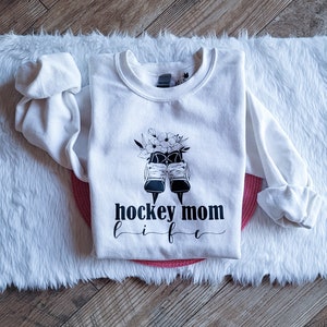 K&ouml;nnte beinhalten: Wei&szlig;er Pullover mit schwarzem Text "hockey mom life" und einer Grafik von Schlittschuhen und Blumen.