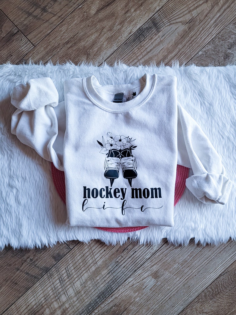 K&ouml;nnte beinhalten: Wei&szlig;er Pullover mit schwarzem Text "hockey mom life" und einer Grafik von Schlittschuhen und Blumen.