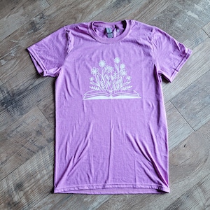 Peut inclure: Un t-shirt violet avec un motif blanc d'un livre ouvert avec des fleurs qui poussent.