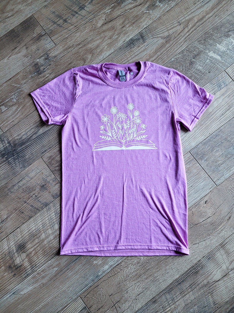 Peut inclure: Un t-shirt violet avec un motif blanc d'un livre ouvert avec des fleurs qui poussent.