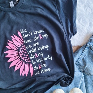 May include: A dark charcoal gray t-shirt featuring a pink sunflower graphic on the left side. The text reads: We don't know how strong we are until being strong is the only choice we have. Three small pink awareness ribbons replace the letter i.