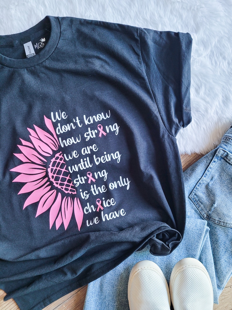 May include: A dark charcoal gray t-shirt featuring a pink sunflower graphic on the left side. The text reads: We don't know how strong we are until being strong is the only choice we have. Three small pink awareness ribbons replace the letter i.