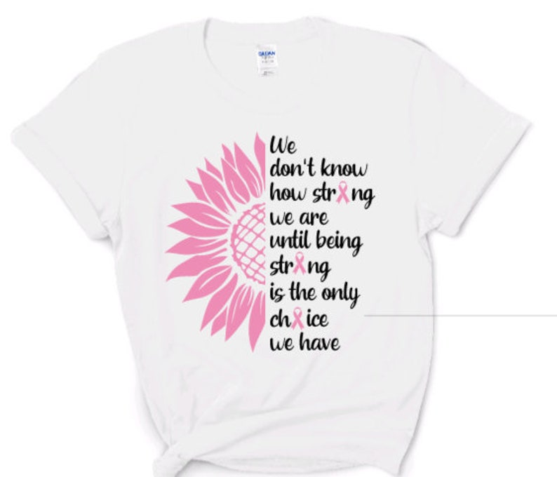 May include: A white short-sleeved t-shirt featuring a graphic of a pink sunflower on the left side and black text that reads: We don't know how strong we are until being strong is the only choice we have, with pink ribbons replacing the letter o.