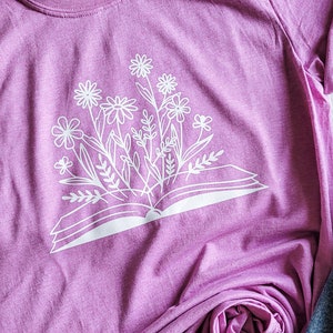 Peut inclure: Un t-shirt rose avec un imprim&eacute; blanc d'un livre ouvert avec des fleurs qui en sortent.