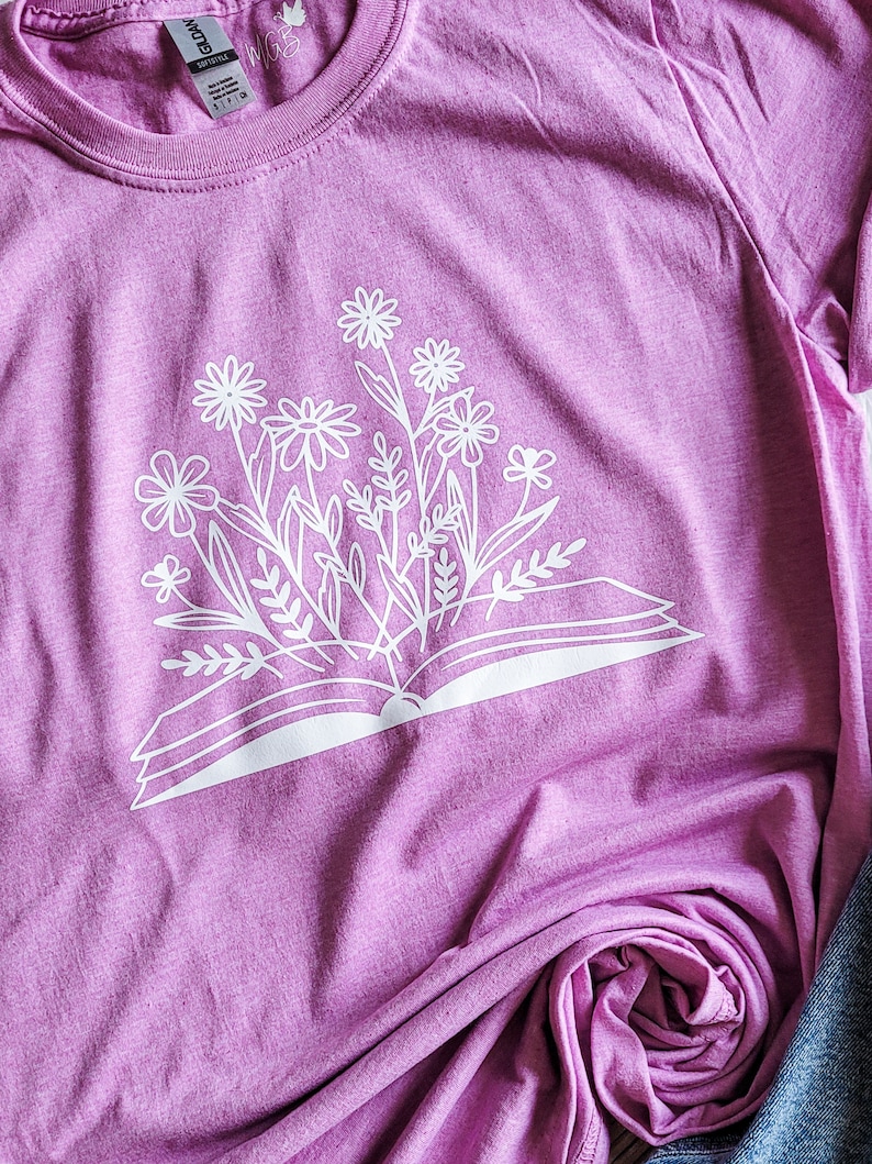 Peut inclure: Un t-shirt rose avec un imprim&eacute; blanc d'un livre ouvert avec des fleurs qui en sortent.