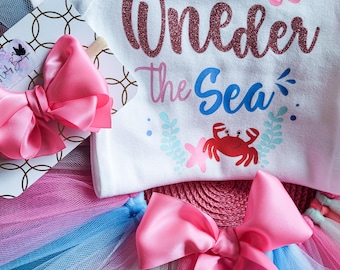 Puede incluir: Un body blanco con el texto "One-der the Sea" en purpurina rosa, con un pulpo rosa y un cangrejo rojo. Se incluye un tutú de tul rosa con capas azules y rosas.
