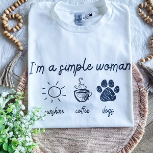 May include: White t-shirt with the text "I'm a simple woman" and three icons: a sun, a coffee cup, and a paw print. The text below the icons reads "sunshine, coffee, dogs".