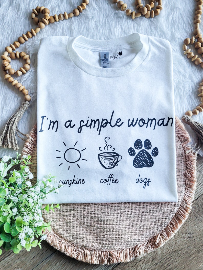 May include: White t-shirt with the text "I'm a simple woman" and three icons: a sun, a coffee cup, and a paw print. The text below the icons reads "sunshine, coffee, dogs".