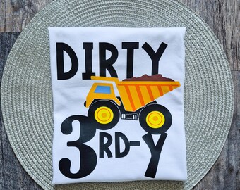 May include: White t-shirt with the words "DIRTY 3RD-Y" in black. A cartoon dump truck in yellow, orange, and brown is printed below the word "DIRTY". The shirt is on a gray placemat and a wooden surface.