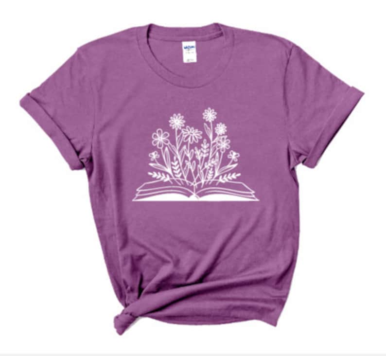 Peut inclure: Un t-shirt violet avec un imprim&eacute; blanc d'un livre ouvert avec des fleurs qui poussent.