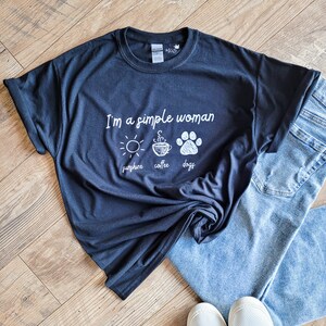 May include: A black t-shirt with white text that reads "I'm a simple woman sunshine coffee dogs". The shirt is laid out on a wood floor with a pair of blue jeans and white sneakers.