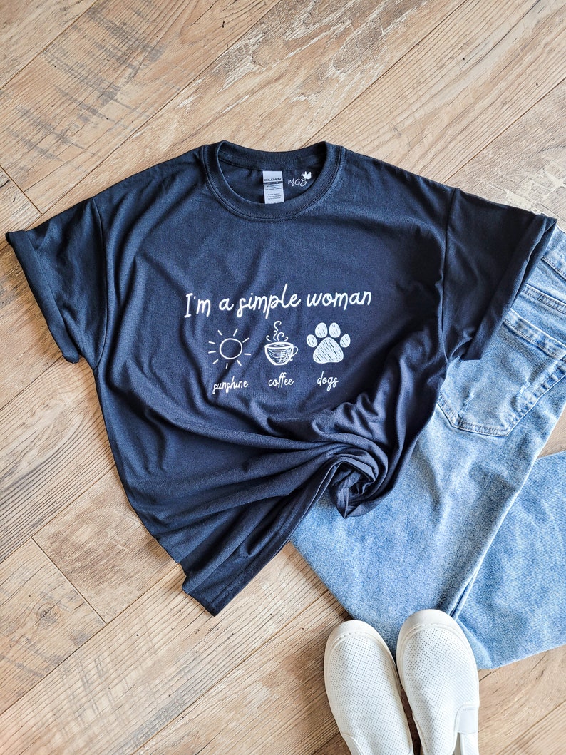 May include: A black t-shirt with white text that reads "I'm a simple woman sunshine coffee dogs". The shirt is laid out on a wood floor with a pair of blue jeans and white sneakers.