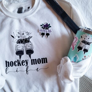 K&ouml;nnte beinhalten: Wei&szlig;es Sweatshirt mit schwarzem Text, der "hockey mom life" lautet, und einer Grafik von Eishockeyschl&auml;gern mit Blumen. Auf dem Sweatshirt befindet sich ein kleinerer Aufkleber mit der Aufschrift "hockey life" und einer Grafik von Eishockeyschl&auml;gern mit Blumen.