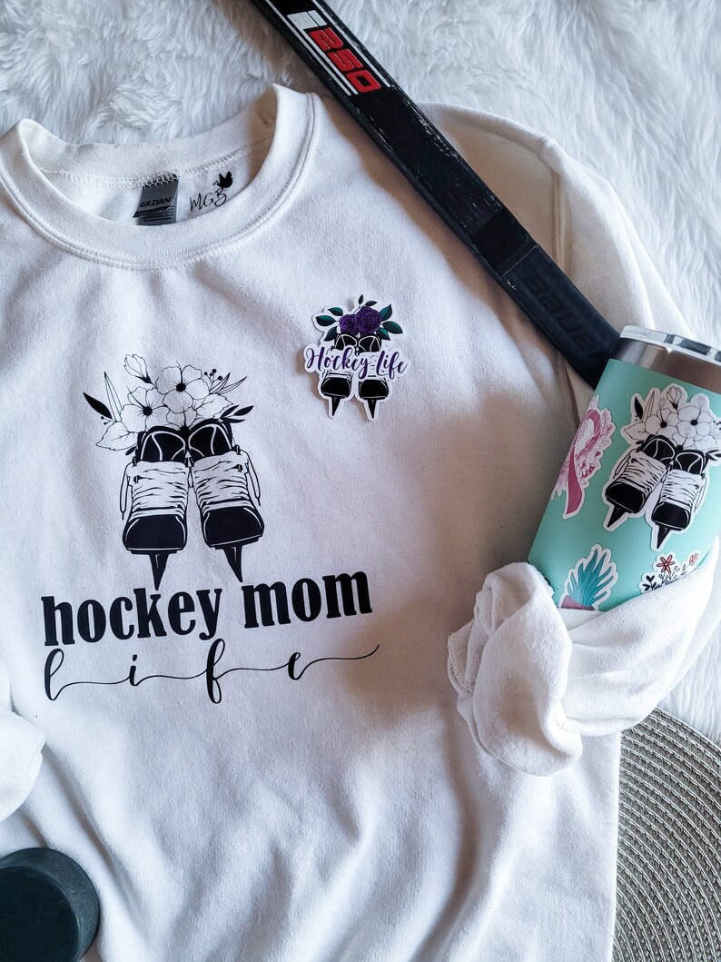 K&ouml;nnte beinhalten: Wei&szlig;es Sweatshirt mit schwarzem Text, der "hockey mom life" lautet, und einer Grafik von Eishockeyschl&auml;gern mit Blumen. Auf dem Sweatshirt befindet sich ein kleinerer Aufkleber mit der Aufschrift "hockey life" und einer Grafik von Eishockeyschl&auml;gern mit Blumen.