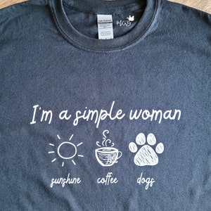 May include: A dark gray t-shirt with white text that reads "I'm a simple woman sunshine coffee dogs". The text is accompanied by three illustrations: a sun, a coffee cup, and a paw print.