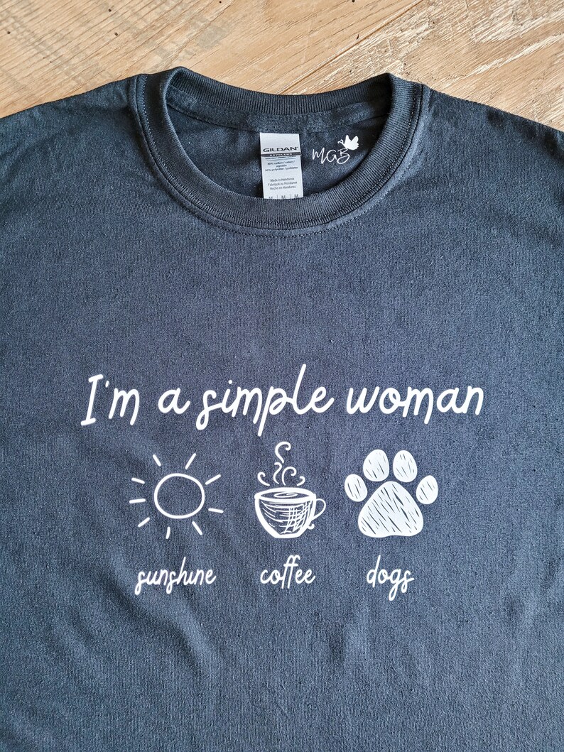 May include: A dark gray t-shirt with white text that reads "I'm a simple woman sunshine coffee dogs". The text is accompanied by three illustrations: a sun, a coffee cup, and a paw print.