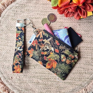 May include: A black fabric zippered pouch and matching wristlet strap featuring a colourful autumn leaf and acorn pattern. The pouch contains a pink lipstick, folded paper currency, and a black card. A small white tag is attached to the zipper.