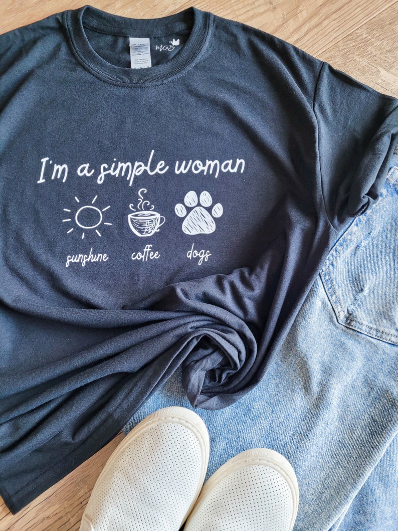 May include: A black t-shirt with the text "I'm a simple woman" and three icons: a sun, a cup of coffee, and a paw print. The icons are labeled "sunshine", "coffee", and "dogs".