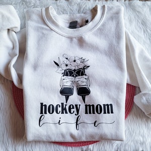 K&ouml;nnte beinhalten: Wei&szlig;er Pullover mit einer schwarz-wei&szlig;en Grafik von Eishockeyschl&auml;gern mit Blumen und dem Text "hockey mom life" darauf gedruckt.