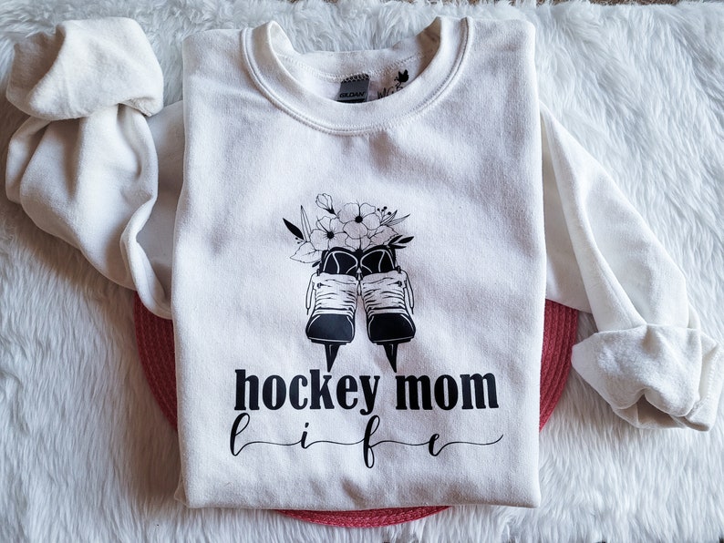 K&ouml;nnte beinhalten: Wei&szlig;er Pullover mit einer schwarz-wei&szlig;en Grafik von Eishockeyschl&auml;gern mit Blumen und dem Text "hockey mom life" darauf gedruckt.