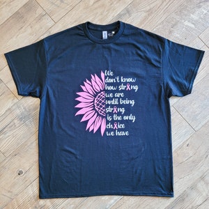 May include: A dark navy blue short-sleeve t-shirt featuring a pink sunflower graphic on the left side. The text reads: We don't know how strong we are until being strong is the only choice we have, with pink awareness ribbons replacing letters.