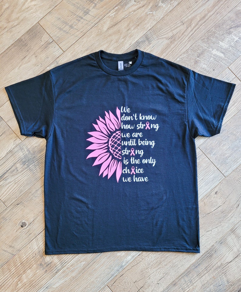 May include: A dark navy blue short-sleeve t-shirt featuring a pink sunflower graphic on the left side. The text reads: We don't know how strong we are until being strong is the only choice we have, with pink awareness ribbons replacing letters.