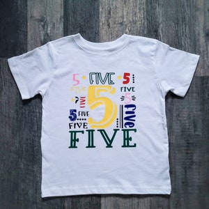 May include: White short-sleeved t-shirt featuring the number "5" in yellow and the word "FIVE" in various colours. The design includes the number 5 and the word "FIVE" in pink, red, blue, and green.