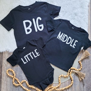 May include: Three black t-shirts and a onesie with white text. The t-shirts read "BIG" and "MIDDLE". The onesie reads "LITTLE". The shirts are arranged on a wooden surface with decorative beads and a white fluffy rug.