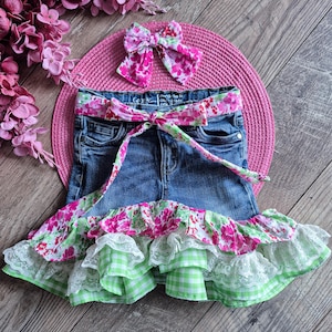 May include: A pair of blue denim shorts featuring a floral print fabric belt, a matching hair bow, and a multi-layered hem with floral print fabric, green and white gingham fabric, and white lace trim, all arranged on a pink circular mat.