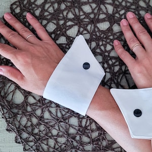 Peut inclure: Une paire de poignets de chemise blancs et rigides, dotés d'une forme pointue et angulaire. Chaque poignet est fixé par un bouton noir, circulaire et brillant. Le tissu semble lisse et structuré, créant un contraste net avec le fond tissé.