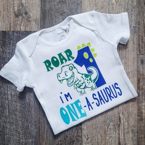 May include: White baby bodysuit with a green and blue dinosaur design. The text reads "ROAR I'M ONE-A-SAURUS" in green and blue. The number 1 is blue with green accents. The bodysuit is 18 months.