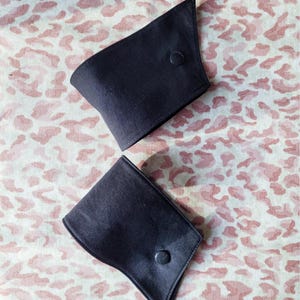 May include: Two dark navy blue cufflink sleeves with a button closure. The cuffs are made of a smooth fabric and are displayed on a pink and white leopard print background.