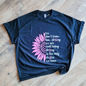 May include: A black short-sleeve t-shirt featuring a pink sunflower graphic on the left side and white text that reads: We don't know how strong we are until being strong is the only choice we have, with pink awareness ribbons replacing letters.