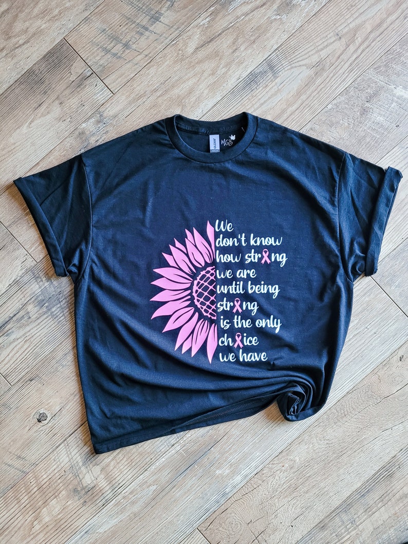 May include: A black short-sleeve t-shirt featuring a pink sunflower graphic on the left side and white text that reads: We don't know how strong we are until being strong is the only choice we have, with pink awareness ribbons replacing letters.