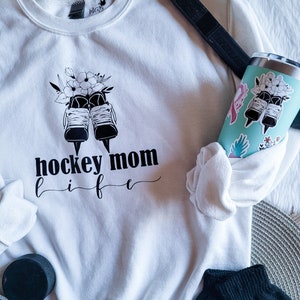K&ouml;nnte beinhalten: Wei&szlig;es Sweatshirt mit einer schwarzen Grafik von Eishockeyschl&auml;gern mit Blumen und dem Text "hockey mom life".