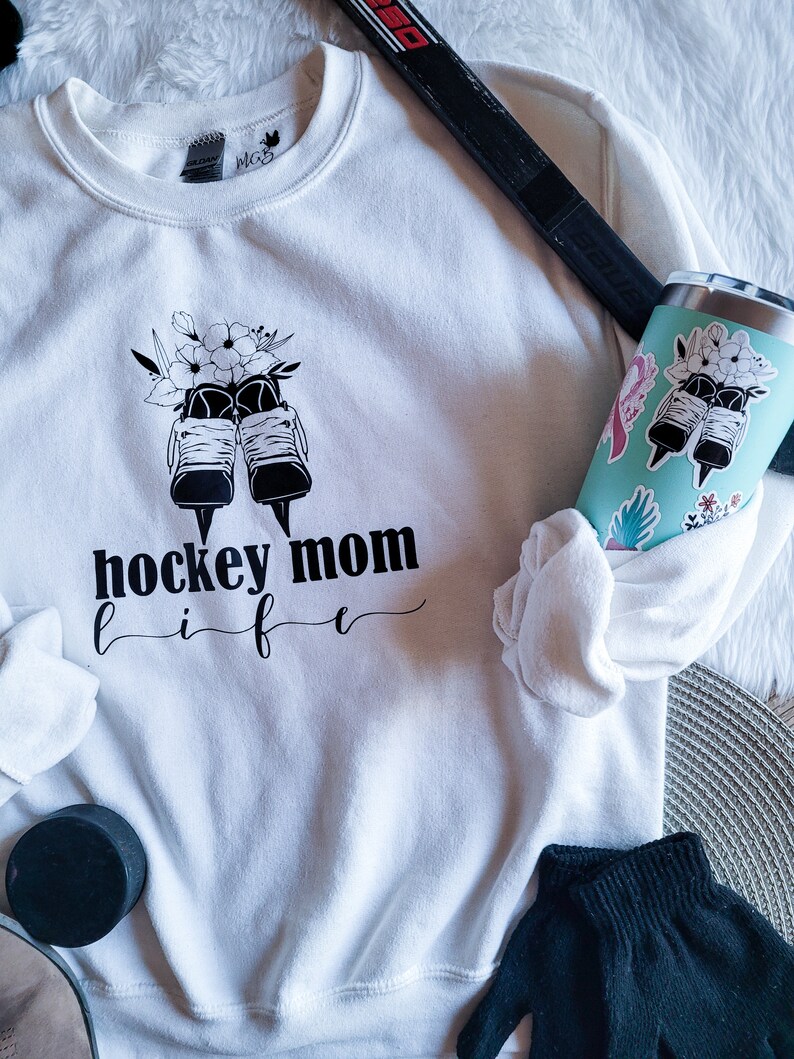 K&ouml;nnte beinhalten: Wei&szlig;es Sweatshirt mit einer schwarzen Grafik von Eishockeyschl&auml;gern mit Blumen und dem Text "hockey mom life".
