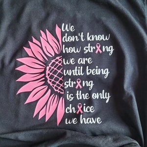 May include: A black t-shirt featuring a pink sunflower graphic on the left side and white text that reads: We don't know how strong we are until being strong is the only choice we have. The letters o in strong and choice are replaced by pink ribbons.
