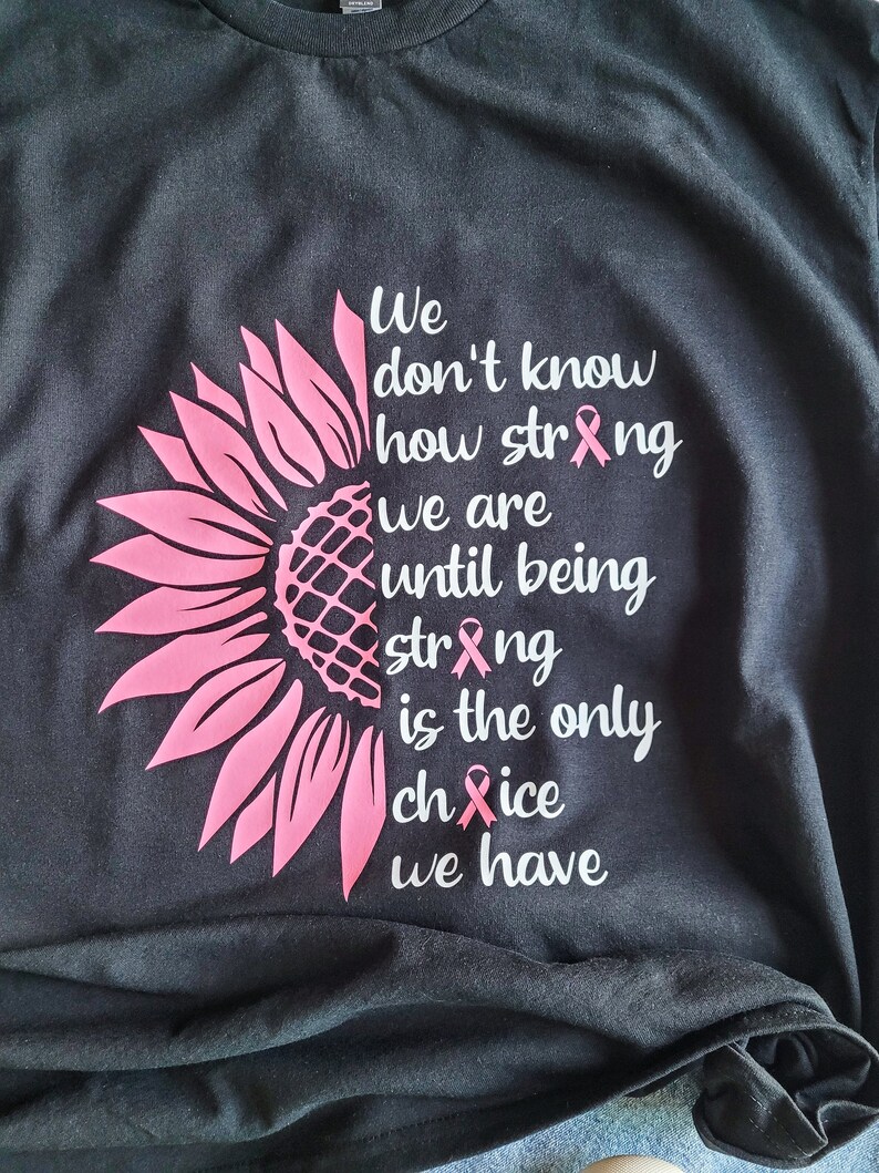 May include: A black t-shirt featuring a pink sunflower graphic on the left side and white text that reads: We don't know how strong we are until being strong is the only choice we have. The letters o in strong and choice are replaced by pink ribbons.