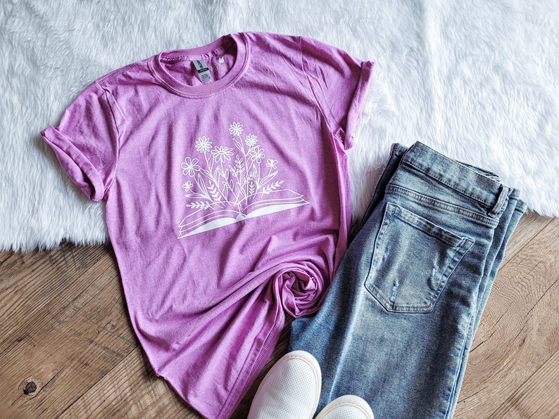 Peut inclure: Un t-shirt rose avec un imprim&eacute; blanc d'un livre ouvert avec des fleurs qui en sortent.