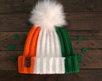 Puede incluir: Un gorro de punto con un pompón blanco, rayas naranja, blanco y verde, y una etiqueta de cuero marrón que dice "Made with Love & Cozy".