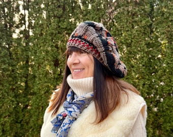 May include: A brown, black, and red crocheted beret. The hat is worn with a cream-colored turtleneck sweater, a blue and white scarf, and a cream-colored vest. The background is a green, leafy tree.