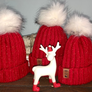 May include: Three red knitted hats with white pom-poms and a brown leather tag that says "Knitted & Sewn". The hats are arranged in a row on a wooden surface with a white ceramic reindeer figurine in the middle.