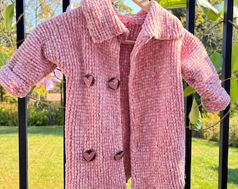May include: A pink knitted cardigan with a collar and heart-shaped buttons. The cardigan is hanging on a wooden hanger.