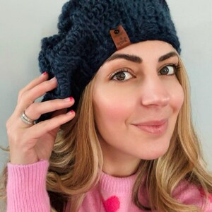 May include: A dark blue knitted beanie with a small brown leather tag. The beanie is made of a thick, textured yarn. The woman in the image is wearing a pink sweater with heart-shaped designs.