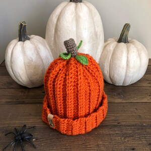 May include: An orange crocheted pumpkin hat with a green stem and brown leaves. The hat is sitting on a wooden surface with three white pumpkins in the background.