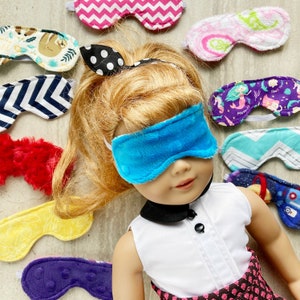 May include: A doll wearing a blue sleep mask with a variety of colorful sleep masks around it. The sleep masks have different patterns, including zigzags, sloths, mermaids, and space themes. The doll has blonde hair and is wearing a white shirt.
