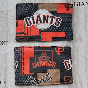 May include: A rectangular fabric patch featuring the San Francisco Giants baseball team logo. The front displays a baseball graphic with the word GIANTS, while the back shows a Golden Gate Bridge illustration and the word GIANTS in orange and black.
