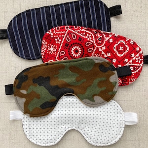 May include: Four different sleep masks. One is black and white striped, one is red with a paisley pattern, one is camouflage, and one is white with a small white dot pattern.